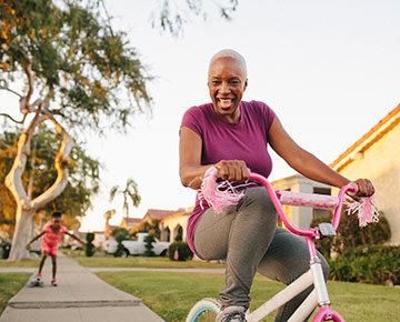 Grandmother riding a child's bike while her granddaughter rides behind her on a skateboard